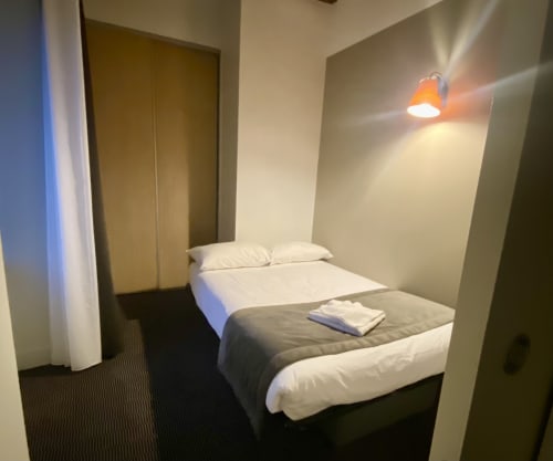 Chambre compacte de l'Hôtel Saint Roch avec lit double, oreillers blancs, moquette foncée, applique murale et murs beige
