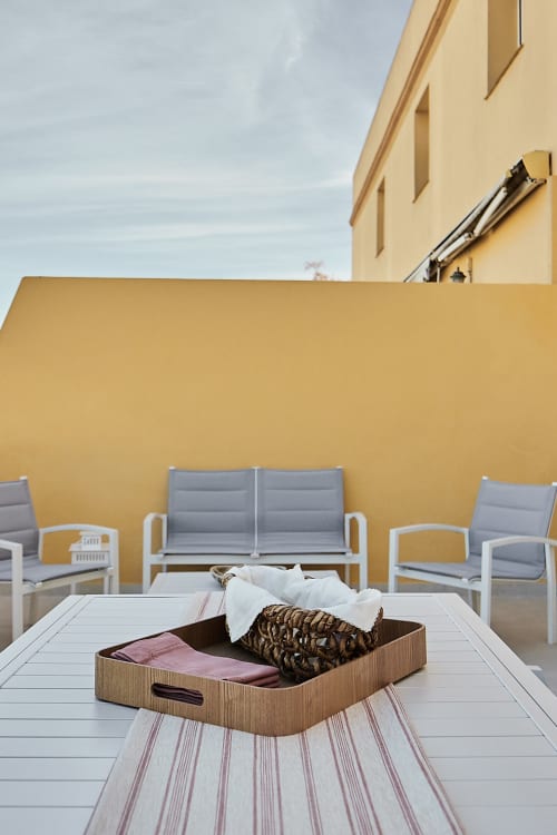 Balcón de apartamento frente al mar en Wehost Costaluz con canasta de almacenamiento de madera, área de estar gris y edificio amarillo