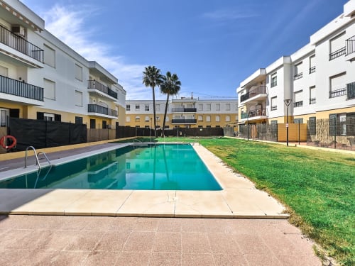 Complejo de apartamentos moderno con piscina turquesa, jardín verde y palmera en Puerto Santa María