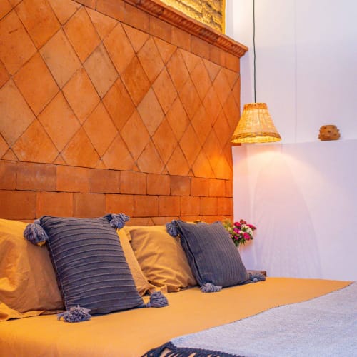 Recámara de Casa Sofi Oaxaca con cabecera de azulejos naranjas, almohadas a rayas azules, ropa de cama amarilla y lámpara colgante tejida.