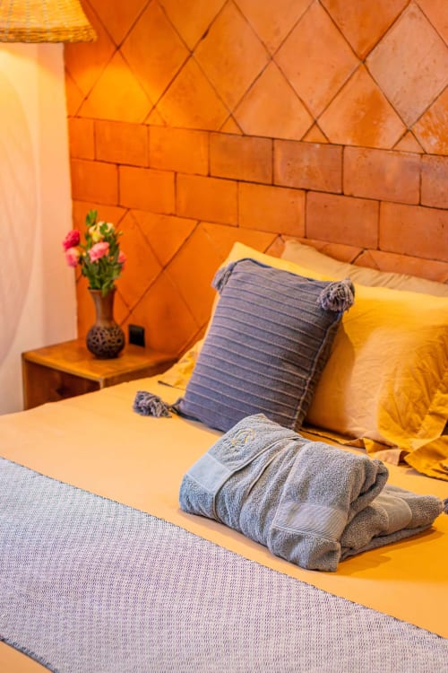 Dormitorio colorido en Casa Sofi Oaxaca con paredes de ladrillo naranja, ropa de cama amarilla, almohada azul y flores rosas en la mesilla.