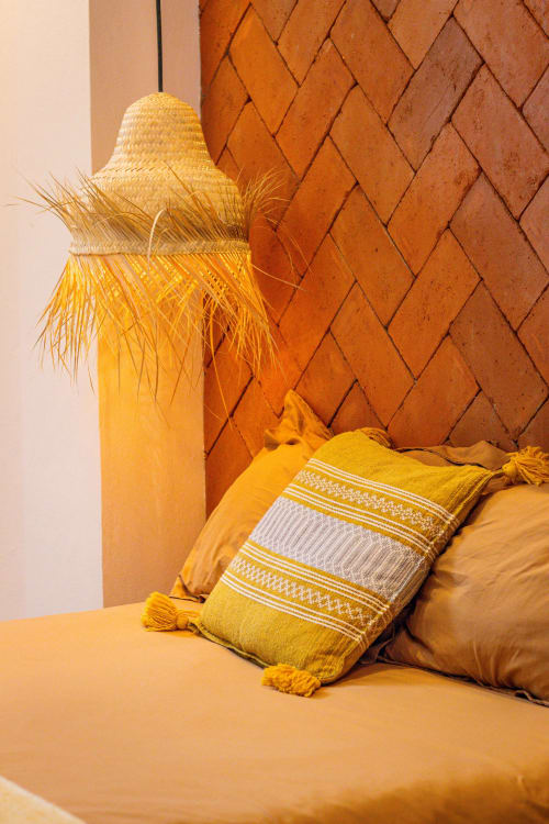 Acogedora habitación en Casa Sofi Oaxaca con ropa de cama dorada, almohada decorativa y sombrero tejido contra pared de ladrillo terracota