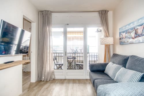 Modern apartment bedroom with balcony view in Palafrugell. Gray sofa, TV, and French doors opening to terrace.