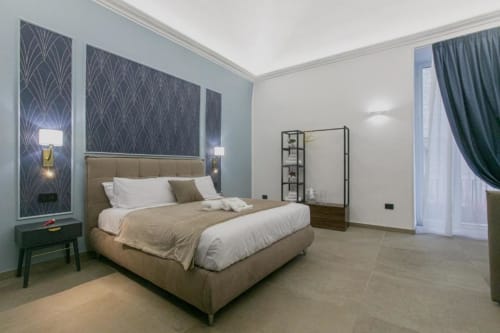 Camera moderna nella Azure Suite di Pignasecca Domus Napoli con letto imbottito beige, parete azzurra e balcone con vista sulla città