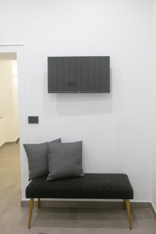 Camera d'albergo moderna con TV a parete sopra divano letto imbottito grigio, gambe in legno, pareti bianche e vista sulla città