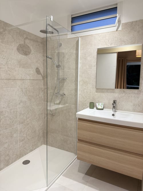 Salle de bains moderne avec douche ouverte, lavabo blanc, meuble vasque en bois et fenêtre bleue au Domaine de la Glacière, Petit Bourg