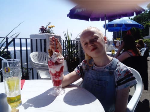 Young child enjoying a red beverage on the outdoor patio at Grouse Lodge in Dunoon, with umbrellas and ocean views visible.