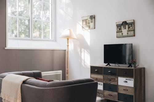 Salon lumineux de La Maison D'olivier avec fauteuil gris, meuble TV, fenêtre vue jardin et lampadaire