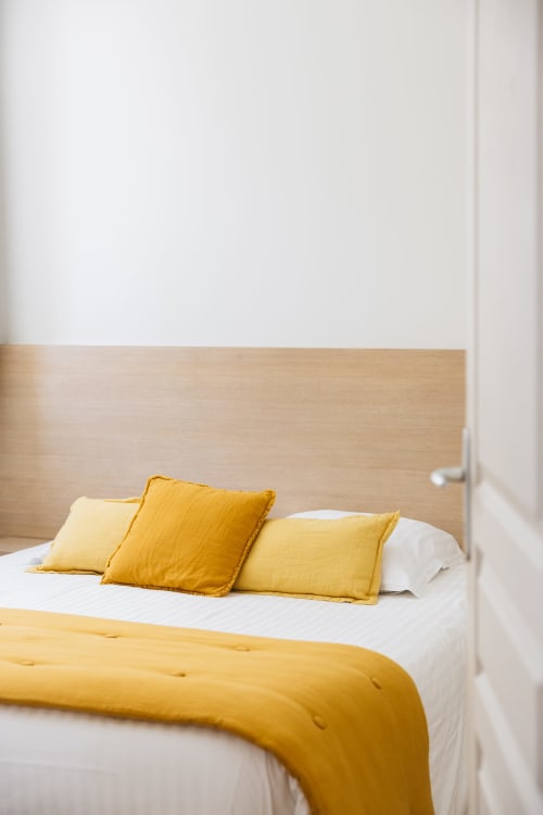 Chambre lumineuse du gîte La Maison D'olivier avec coussins jaunes et plaid sur lit blanc à tête de lit bois