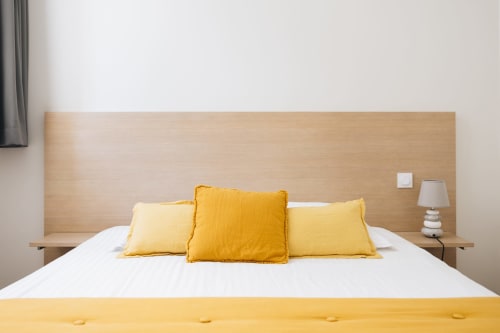 Chambre lumineuse à la maison d'Olivier avec oreillers jaunes, literie blanche, tête de lit en bois et décoration moderne