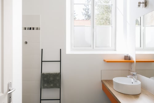 Salle de bain moderne du gîte La Maison D'olivier avec lavabo rond blanc, meuble en bois, étagères orange et échelle à serviettes noire