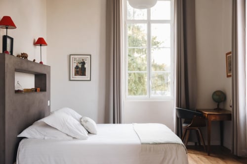 Chambre lumineuse à La Maison D'olivier avec lit blanc, tête de lit grise, lampes rouges et vue sur le jardin à Mesnard la Barotière