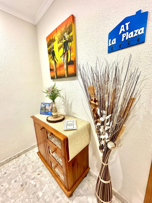 Entrada del departamento en Mérida con mueble de madera, obra de arte al atardecer, plantas decorativas y letrero 'At La Plaza'