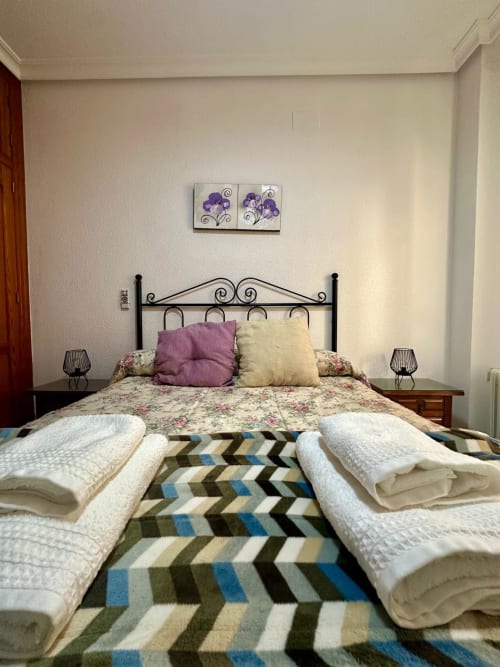 Dormitorio acogedor en apartamento Gestyp-AT en Mérida con cama de hierro forjado, ropa de cama floral, acentos morados y toallas enrolladas
