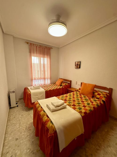 Dormitorio triple con colchas rojo y amarillo, cortinas naranjas y ventilador de techo moderno en apartamento de Mérida