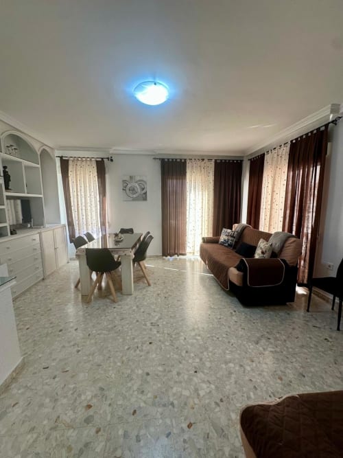 Amplia sala de estar moderna en Mérida con sofá marrón, comedor, estantería blanca y grandes ventanales con marcos marrones