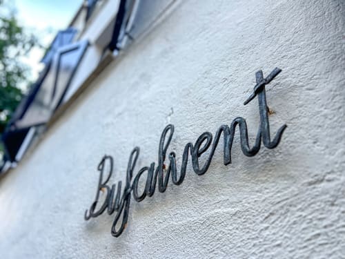Metal 'Bulganent' sign with cross on white textured wall at Finques Garcia Enrich, Palafrugell