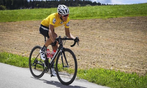 Cyclist in yellow jersey riding on rural road near Calamandrana with green fields and forest backdrop