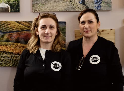 Due dipendenti di Agorà Calamandrana in uniforme nera con loghi, in piedi davanti a un'opera d'arte con vigneto.