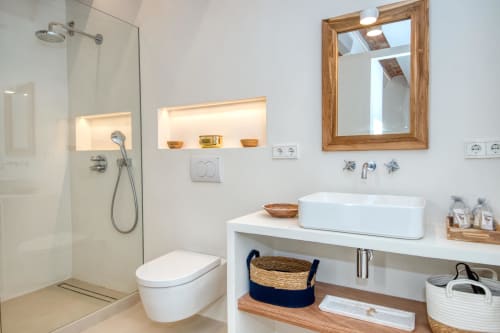 Modern bathroom in Palafrugell cottage with white vessel sink, wooden mirror, rainfall shower, and wooden storage baskets