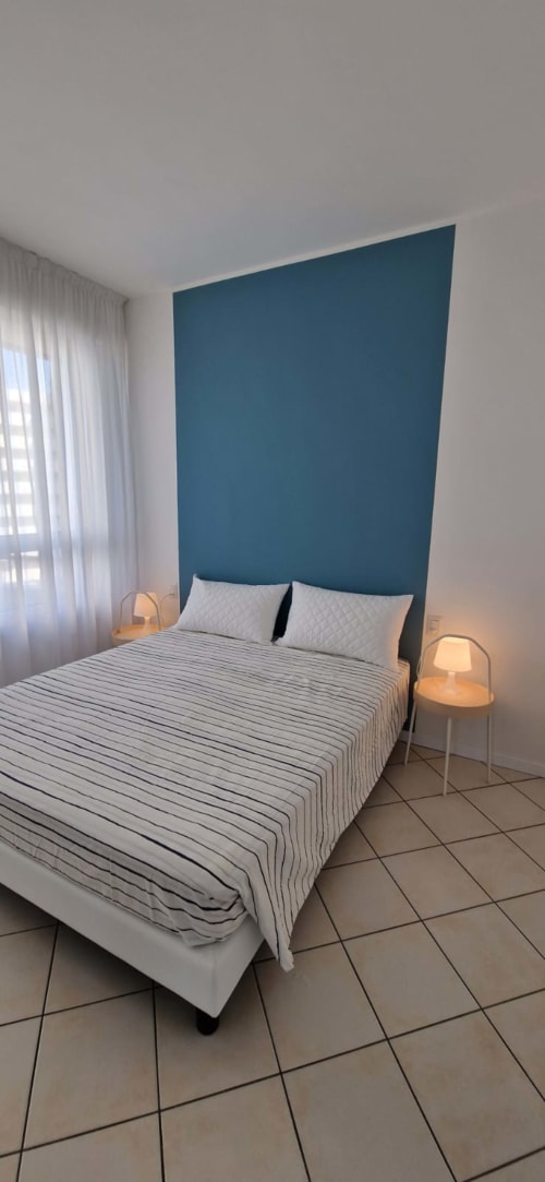 Modern bedroom with teal accent wall, striped bedding, side lamps, sheer curtains, and tile flooring at Lamar Suites Seafront