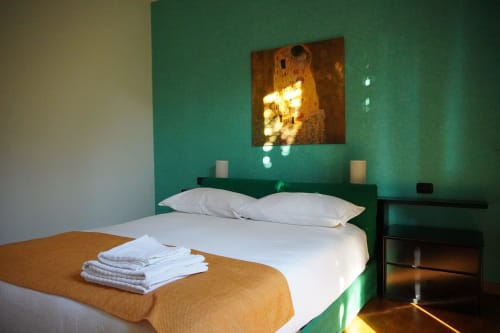 Roma 1 Apartment bedroom with turquoise wall, white bed, orange blanket, and golden artwork at THE V Collection in Turate,