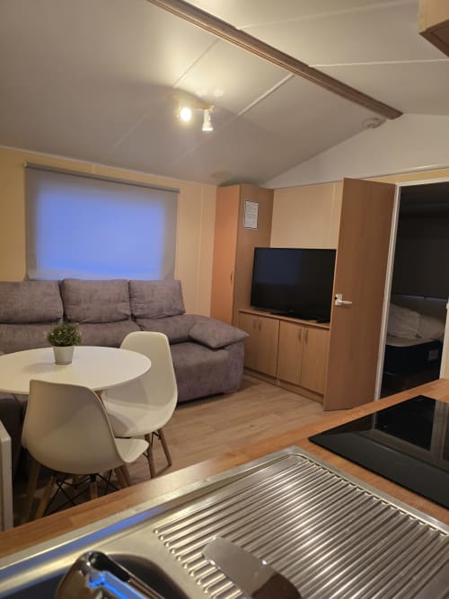 Modern bungalow living area with gray sofa, white dining table, TV, and wood furnishings in Chipiona, Spain