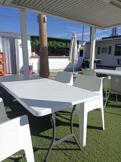 Outdoor dining area at Bungalow Buda Beach in Chipiona with white tables, chairs, and patio umbrella under pergola structure.