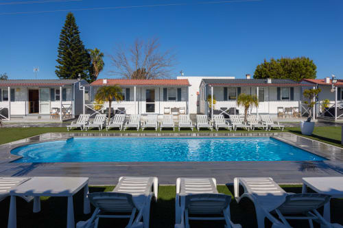 Blue swimming pool with white lounge chairs at Bungalow Buda Beach hotel in Chipiona, Spain, with bungalows and gardens