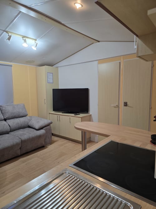 Bungalow G living area at Buda Beach Chipiona with gray sofa, flat-screen TV, wooden furniture and kitchen counter