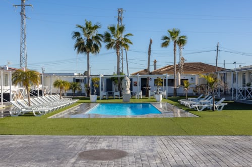 Bungalow Buda Beach pool courtyard in Chipiona with loungers, palm trees, and whitewashed bungalows under clear sky