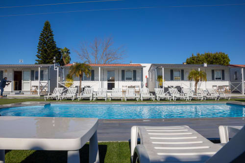 Área de piscina Bungalow Buda Beach en Chipiona con tumbonas, bungalows blancos y palmeras bajo cielo despejado