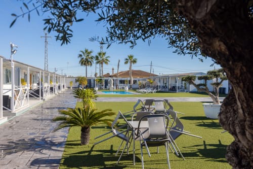 Jardín y patio de Bungalow Buda Beach con sillas de comedor, piscina, palmeras y bungalows blancos en Chipiona
