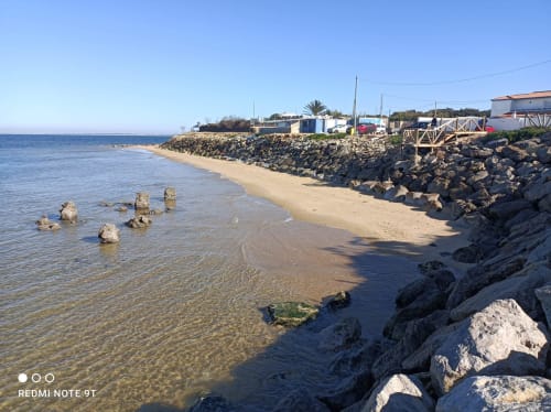 Rocky beach cove in Chipiona with sandy shore, calm blue waters, and beachfront bungalows under clear sky