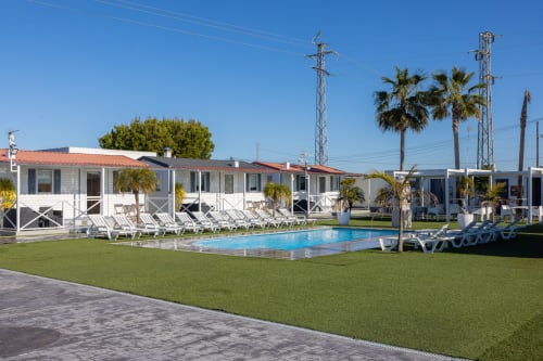 Bungalow Buda Beach resort in Chipiona with white bungalows, blue swimming pool, palm trees, and green lawn under clear sky