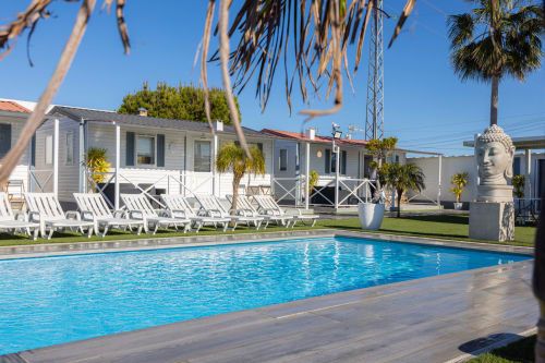 Bungalow Buda Beach pool area in Chipiona with loungers, palm trees, and white bungalows under blue sky