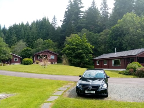 Black Mercedes parked at Grouse Lodge in Dunoon with red chalets and dense Scottish forest backdrop