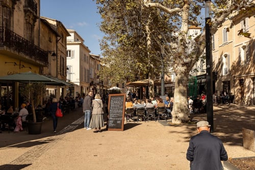 Charmante place ombragée à Montfrin avec terrasse de café et bâtiments historiques, idéale pour les clients du Prieuré