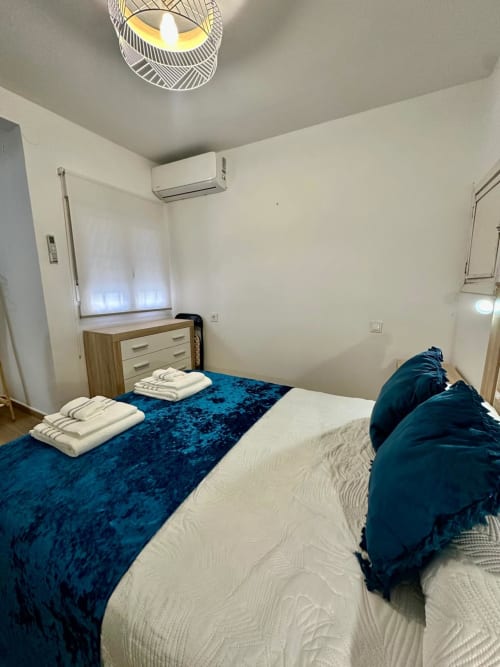 Dormitorio moderno del apartamento Gestyp-AT en Mérida con cama doble, camino azul de terciopelo, cómoda de madera y aire acondicionado de pared