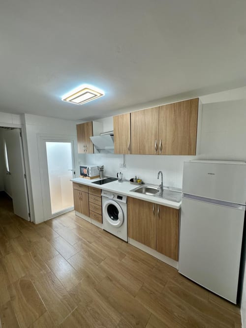 Cocina moderna con muebles de madera, encimeras blancas, electrodomésticos de acero inoxidable y lavadora en apartamento de Mérida