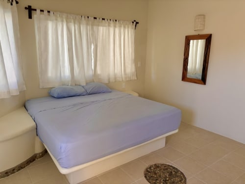 Dormitorio luminoso con cama queen, almohadas azules, cortinas translúcidas, espejo de madera y piso de cerámica en Casa Calilla