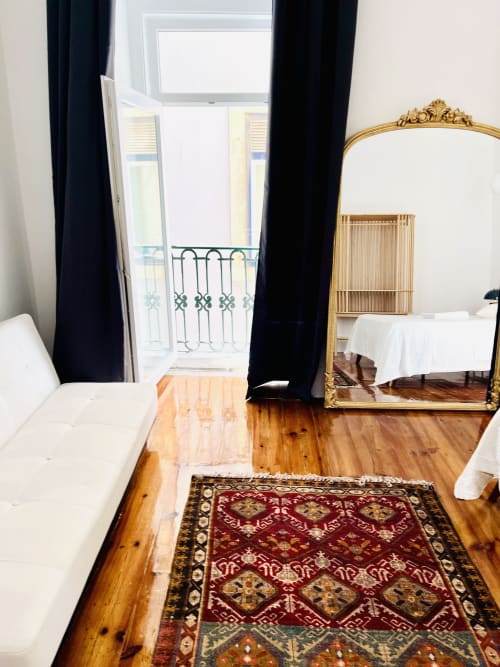 Elegant Lisbon apartment bedroom with wooden floors, ornate gold mirror, black curtains, balcony view, and decorative rug