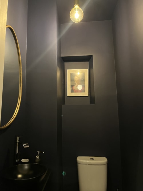 Modern bathroom at Royal Prince Garden Apartments with dark walls, round mirror, white toilet, and framed portrait above in