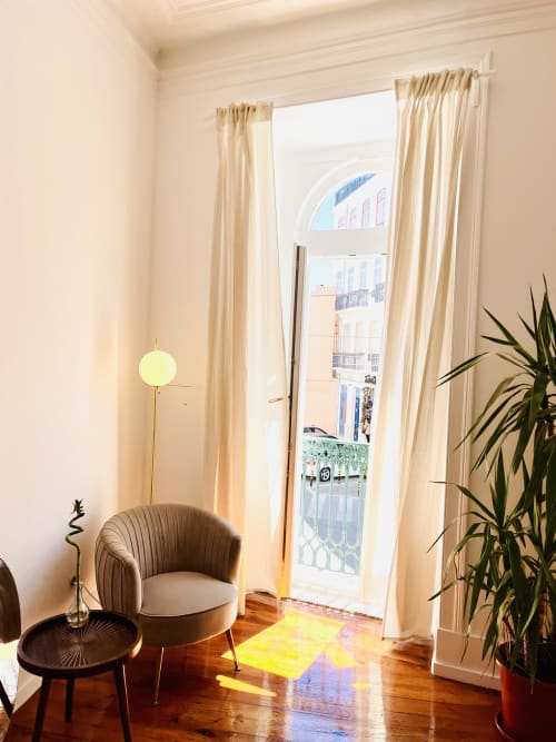 Bright Lisbon apartment living room with cream curtains, wooden floors, modern chair, and city views through glass doors