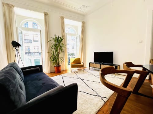 Modern living room in Lisbon apartment with black sofa, wooden chairs, TV, tall windows with city views, potted plant, and