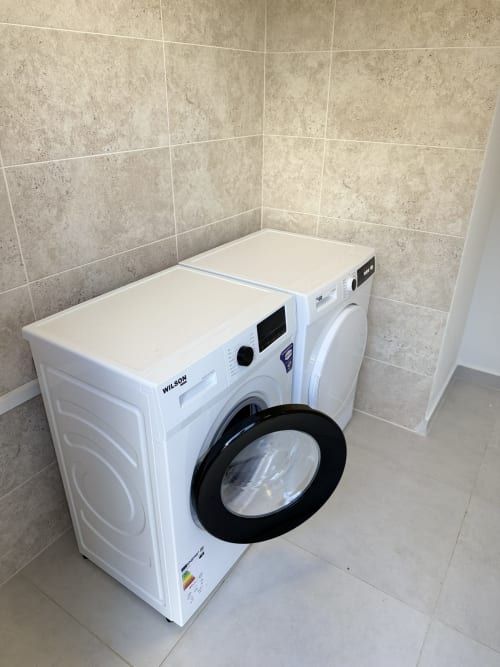 Deux machines à laver Wilson blanches avec hublots noirs dans une buanderie aux murs carrelés beige du Domaine de la
