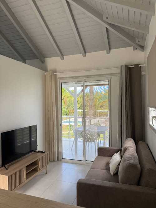 Salon moderne de la Domaine de la Glacière avec plafond à poutres grises, canapé marron, télévision et baies vitrées donnant sur la piscine