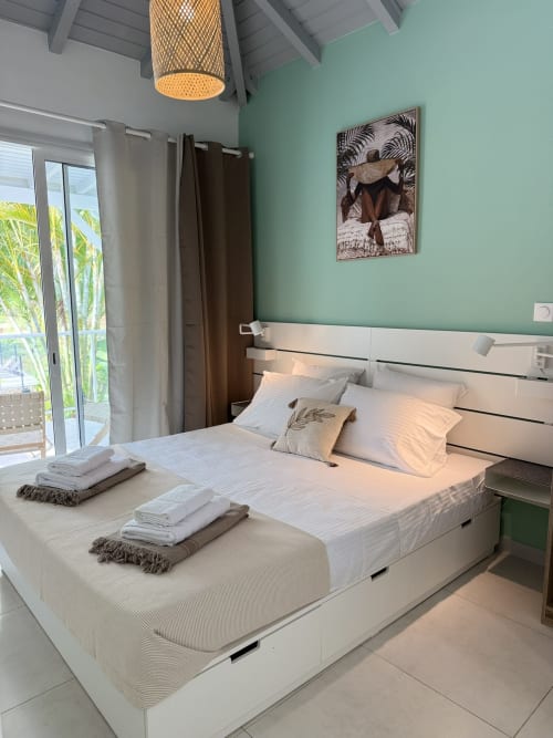 Chambre moderne à la Domaine de la Glacière aux murs vert menthe, lit blanc surélevé, luminaire suspendu tressé et vue sur l'océan