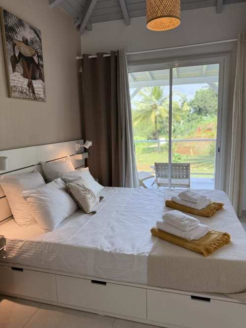 Chambre du bungalow Tiaré au Domaine de la Glacière avec vue sur la piscine, lit blanc, décor tropical et accès à la terrasse.