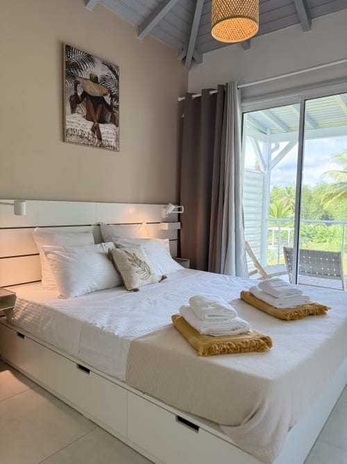 Chambre du bungalow Tiaré au Domaine de la Glacière avec vue sur la piscine, lit blanc et décoration tropicale